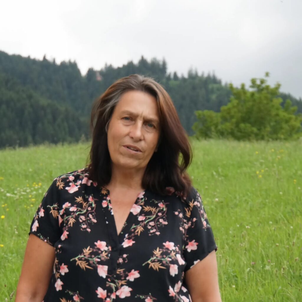 Manuela Högg in einer grünen Sommerwiese mit Blumen, im Hintergrund ein bewaldeter Hügel – Porträt einer Frau in der Natur, Coach für Mensch-Hund-Beziehungen und energetische Begleitung.